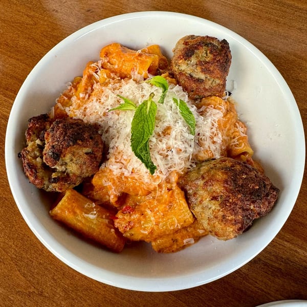 Fratelli By Jholano's rigatoni pasta with orange sauce, meatballs, parmesan, and mint in a white bowl.