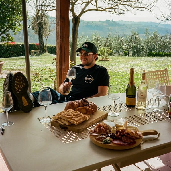 Man relaxing outdoors with wine, Italian charcuterie, and pastries on a table.
