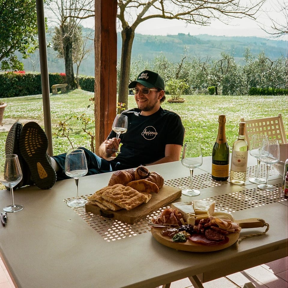 Man relaxing outdoors with wine, Italian charcuterie, and pastries on a table.