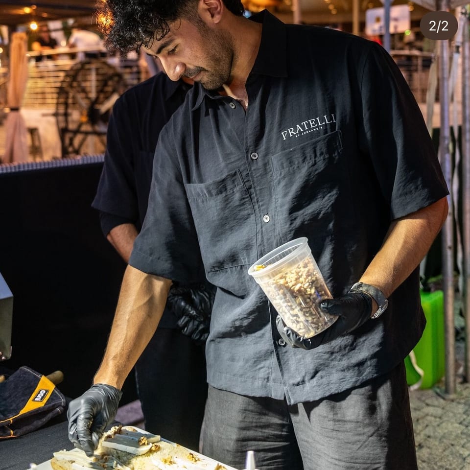 A Fratelli By Jholano's chef in black gloves prepares food, holding a container of nuts.