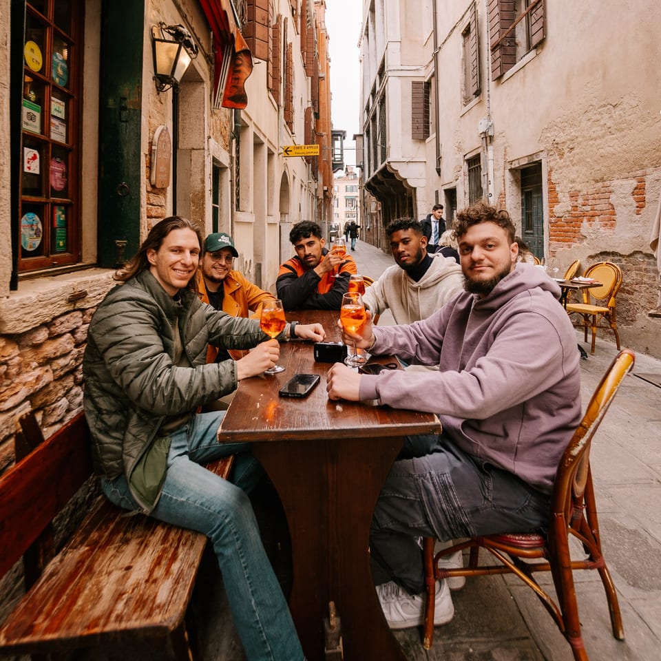 Five men enjoying drinks at an outdoor table in a narrow, historic Italian alleyway.