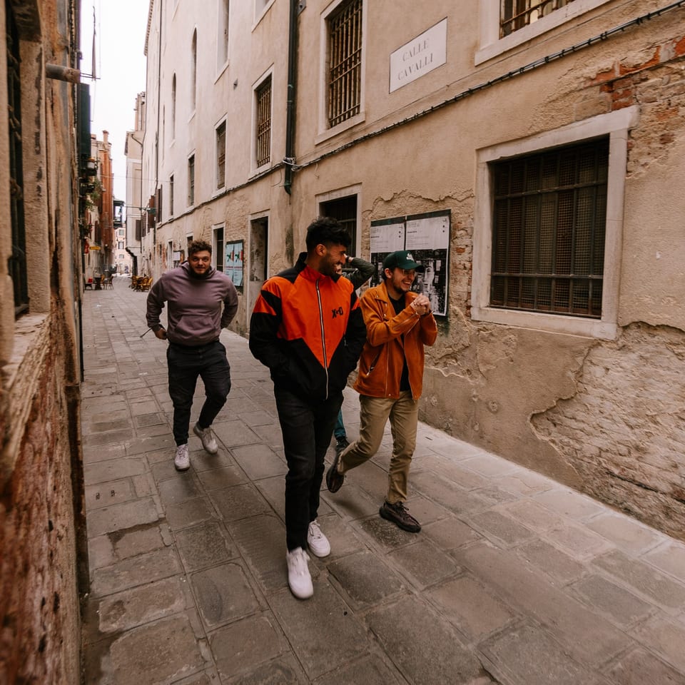 Three men walking and laughing down a narrow, old Italian street with stone buildings.