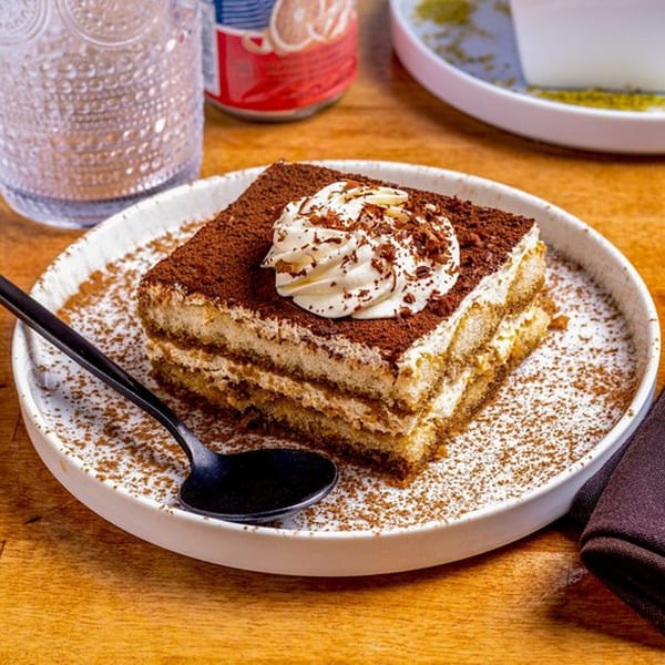 A slice of tiramisu with whipped cream and chocolate shavings on a white plate with a spoon.