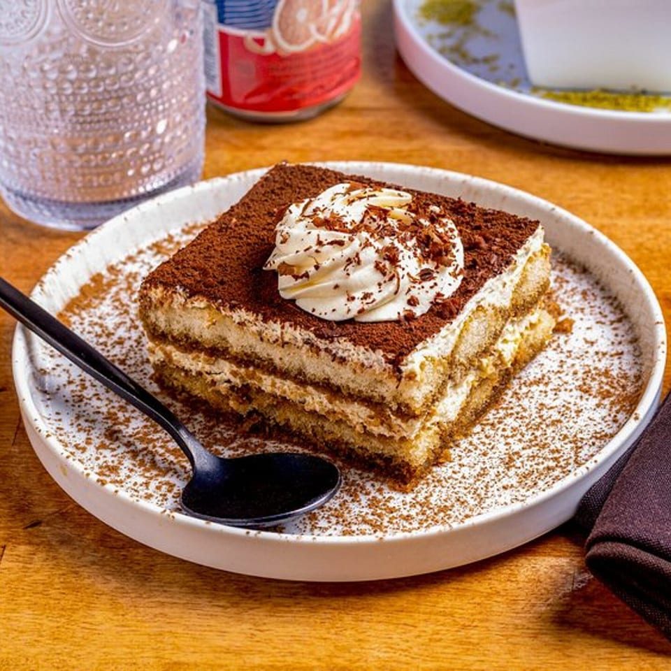 A slice of tiramisu with whipped cream and chocolate shavings on a white plate with a spoon.
