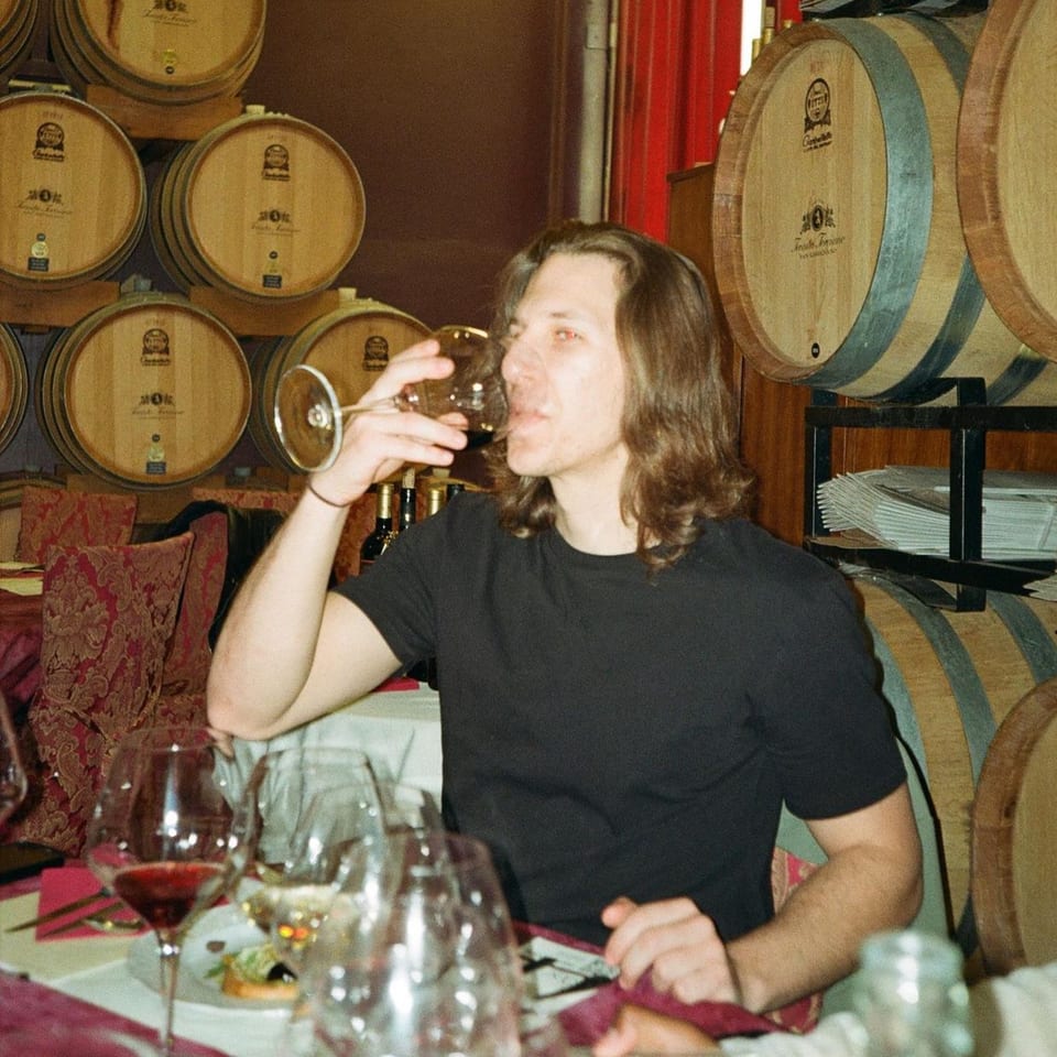 Man with long hair sips wine at a table with wine barrels in the background.