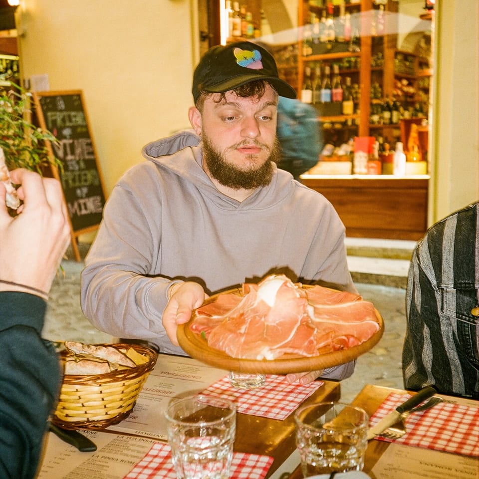 A man in a baseball cap and hoodie holds a wooden platter of prosciutto and burrata at an Italian restaurant.
