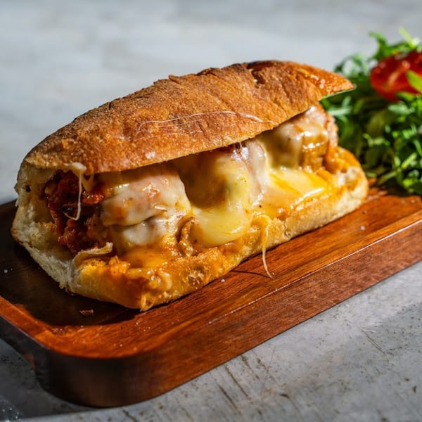 A Fratelli By Jholano's meatball sub with melted cheese and sauce on a wooden board.